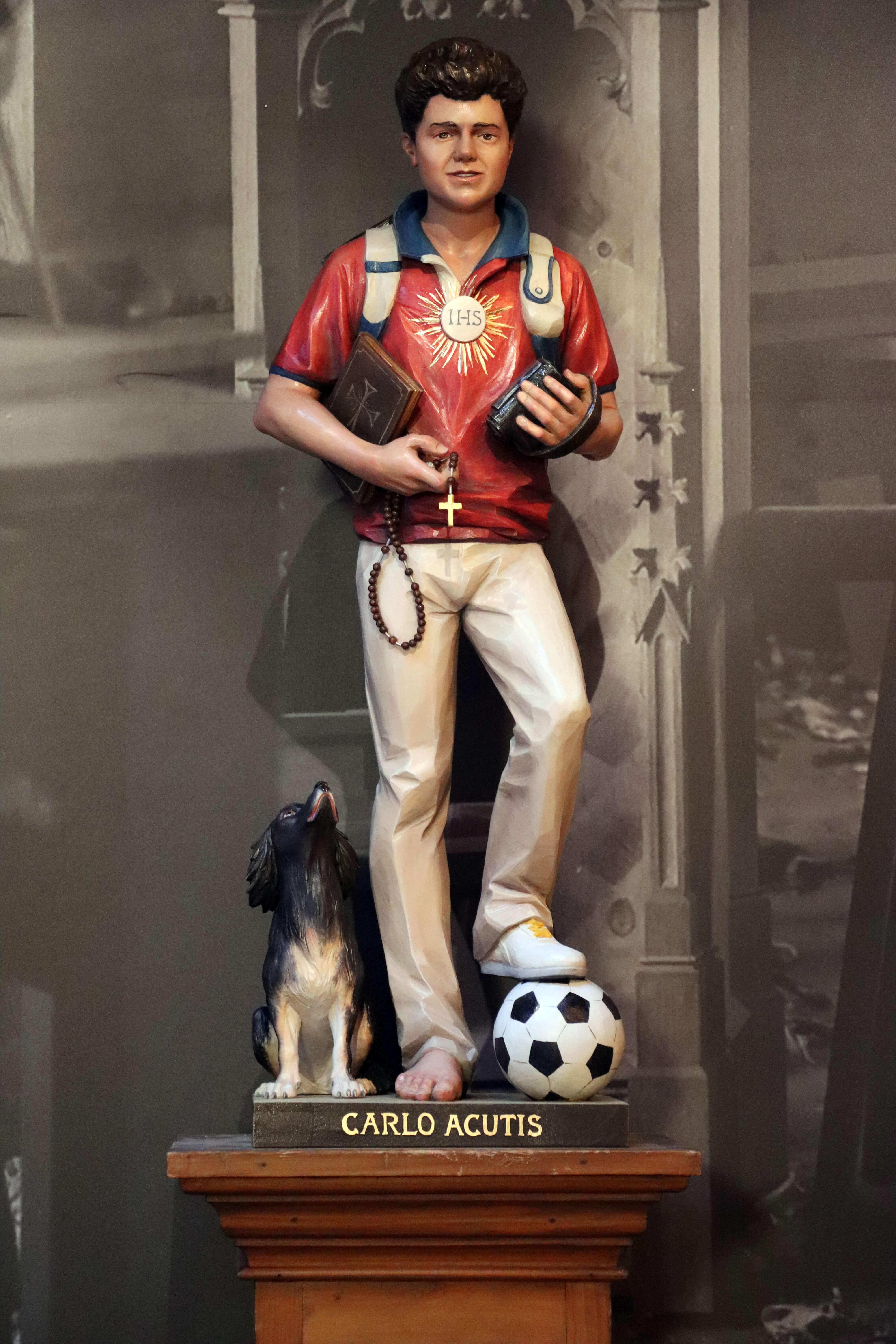 Fertige Statue von Carlo Acutis, die ihn mit einem Rucksack auf dem Rücken zeigt. In einer Hand hält er eine Bibel und einen Rosenkranz, in der anderen einen Camcorder. Ein Hund sitzt am Boden neben ihm und schaut ihn an, während Carlo einen Fuß auf einem Fußball hat. Diese Statue wurde von der Schreinerei Ferdinand Stuflesser unter der Leitung von Robert Stuflesser erschaffen.