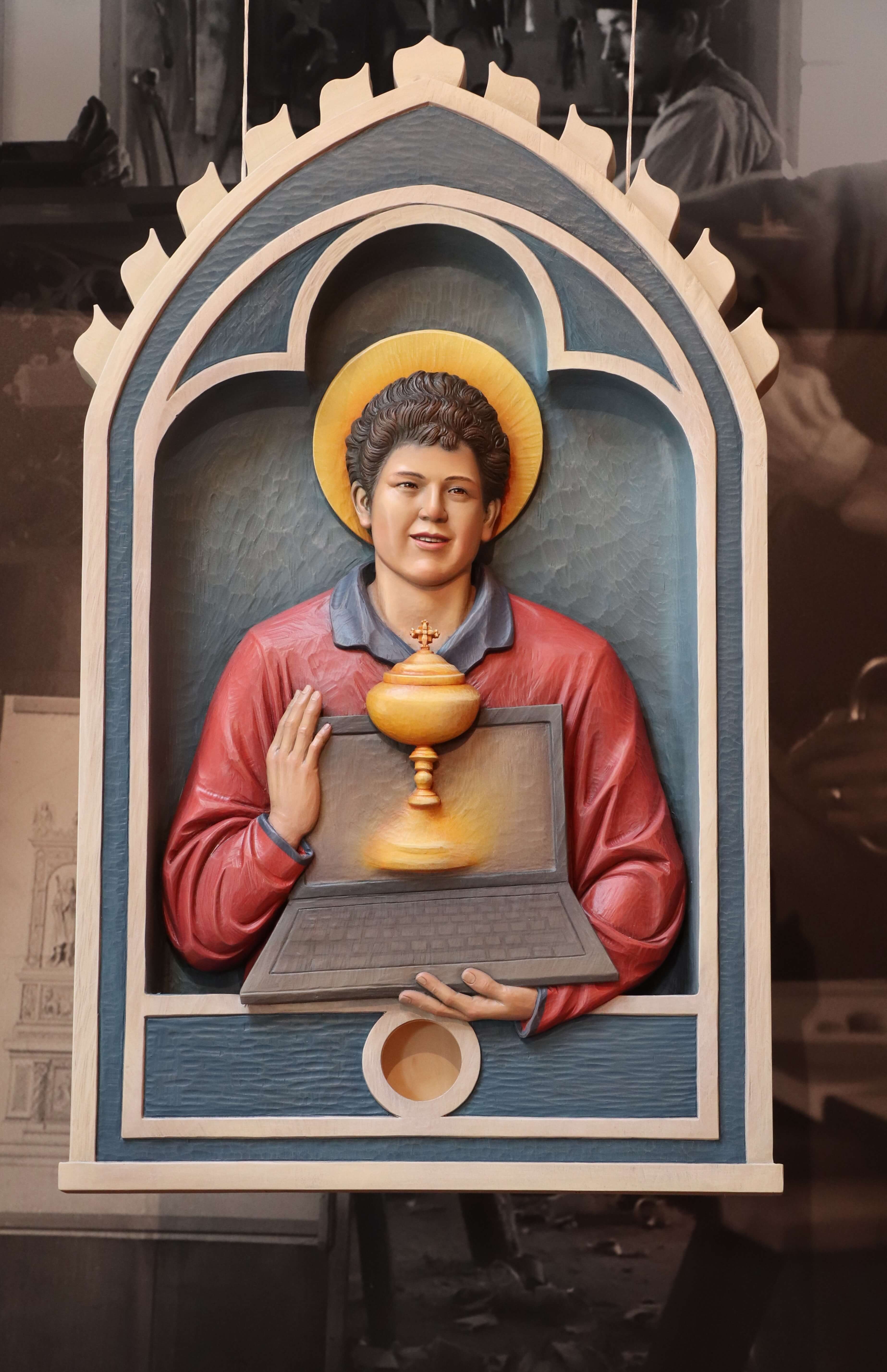 Drei-dimensionales Bild von Carlo Acutis mit einem heiligen Schein über dem Kopf. Er hält einen Laptop, aus dem ein goldener Kelch erscheint. Diese Statue wurde von der Schreinerei Ferdinand Stuflesser unter der Leitung von Robert Stuflesser erschaffen und symbolisiert die Verbindung von Technologie und christlichem Glauben.