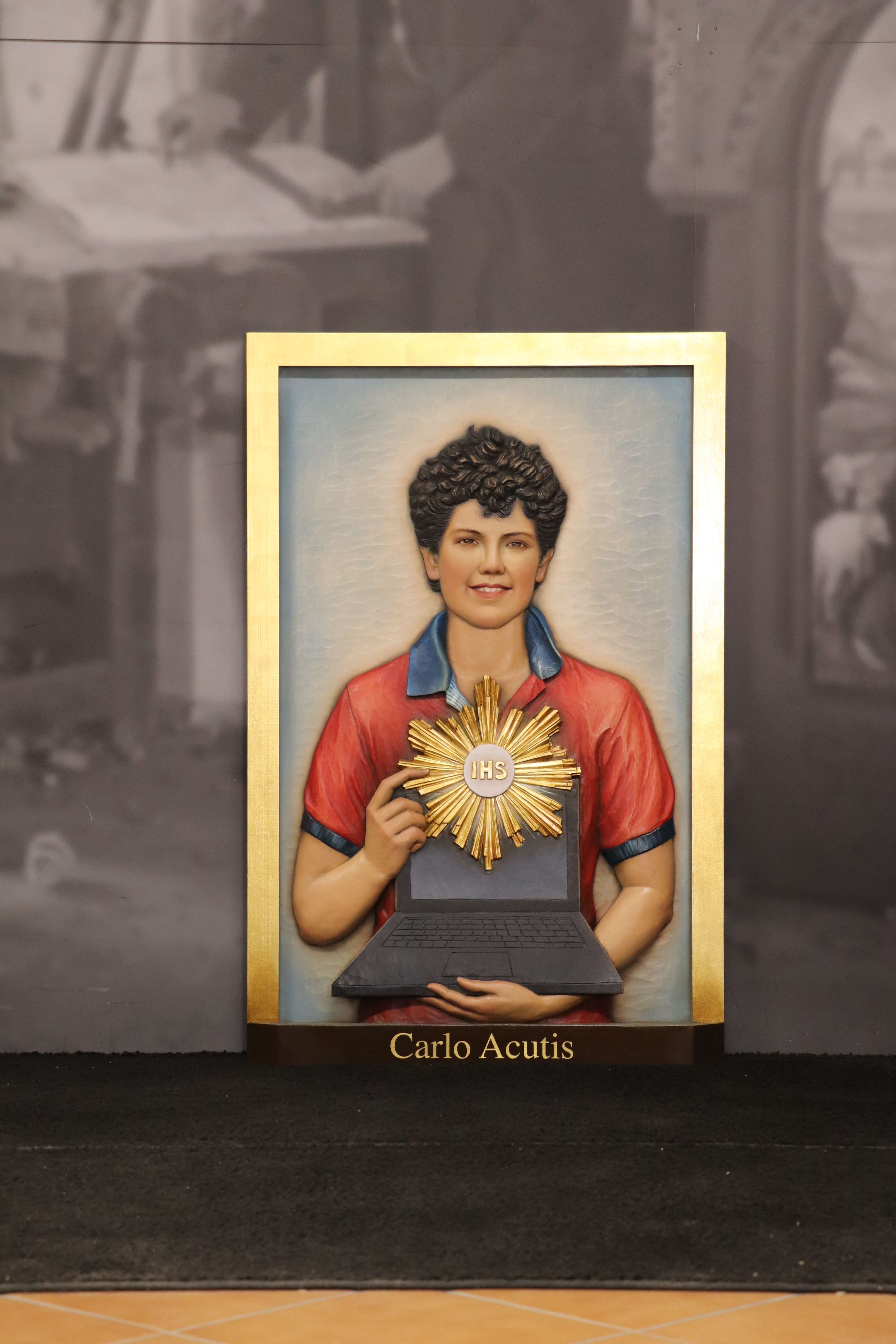 Drei-dimensionales Holzbild von Carlo Acutis, das ihn mit einem Laptop zeigt, auf dessen Bildschirm das christliche Symbol JHS zu sehen ist. Die Statue stellt Carlo in einer modernen Pose dar, die seine Verbindung zwischen Glaube und Technologie widerspiegelt.