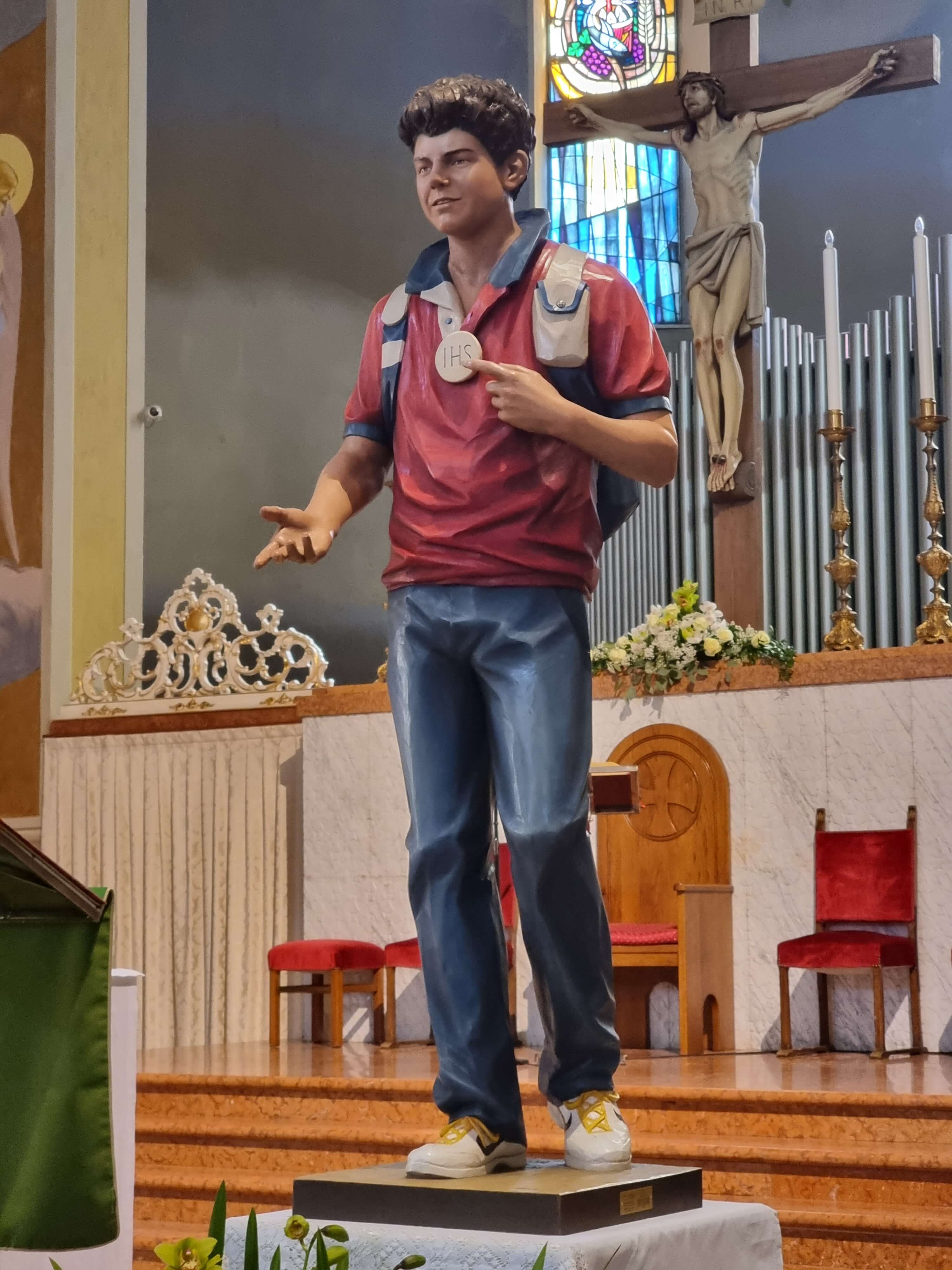 Holzstatue von Carlo Acutis in einer Kirche, die ihn mit einer ausgestreckten Hand zeigt, als würde er sie jemandem reichen. Mit der anderen Hand deutet er auf eine JHS-Kette, die er um seinen Hals trägt. Diese Statue wurde von der Schreinerei Ferdinand Stuflesser unter der Leitung von Robert Stuflesser erschaffen.
