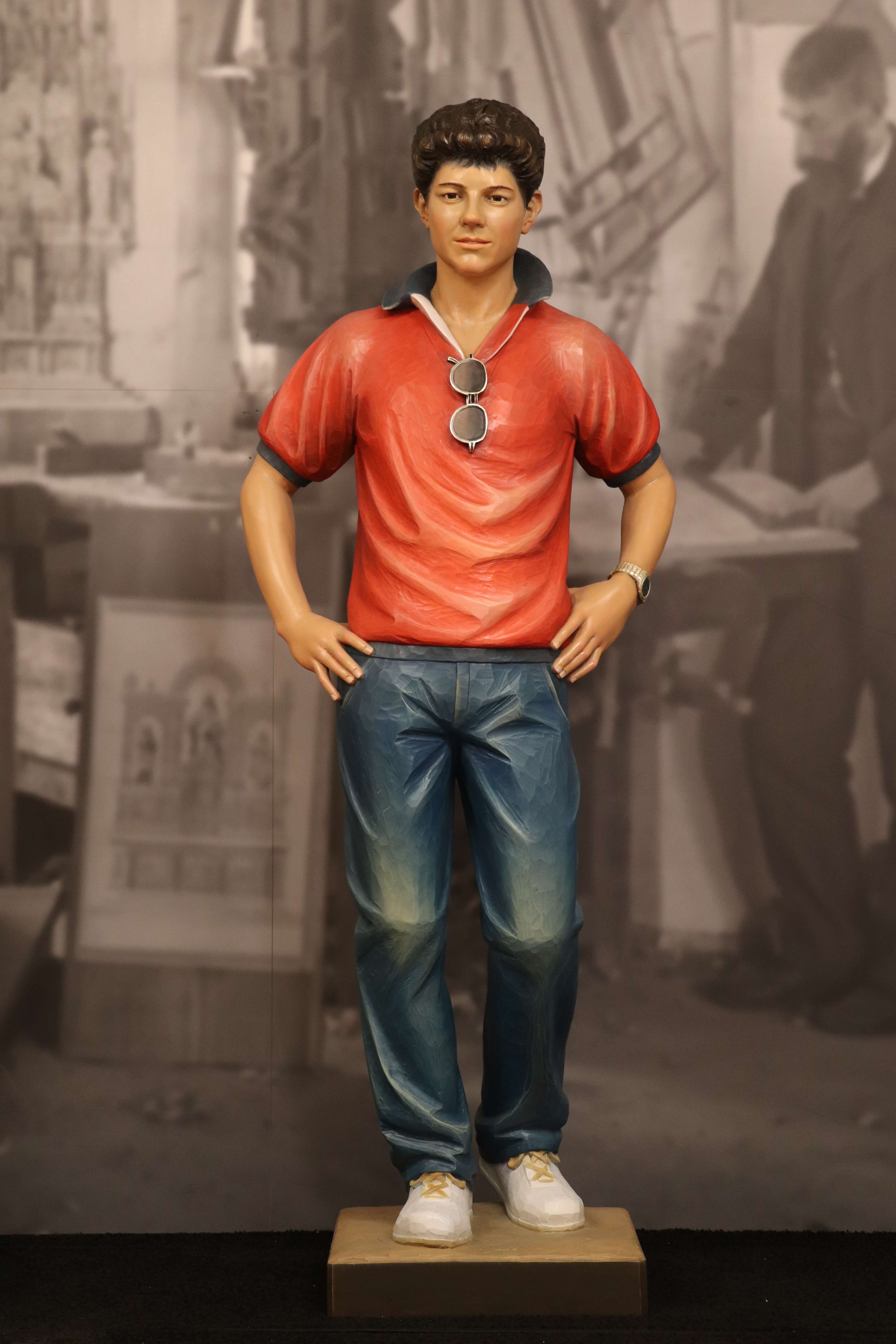 Statue von Carlo Acutis, die ihn in einem roten T-Shirt zeigt. Am Kragen des T-Shirts hängt eine Sonnenbrille. Diese Statue, erschaffen von der Schreinerei Ferdinand Stuflesser unter der Leitung von Robert Stuflesser, stellt Carlo Acutis in einem modernen und jugendlichen Moment dar und kombiniert seine zeitgenössische Ausstrahlung mit seiner spirituellen Hingabe.