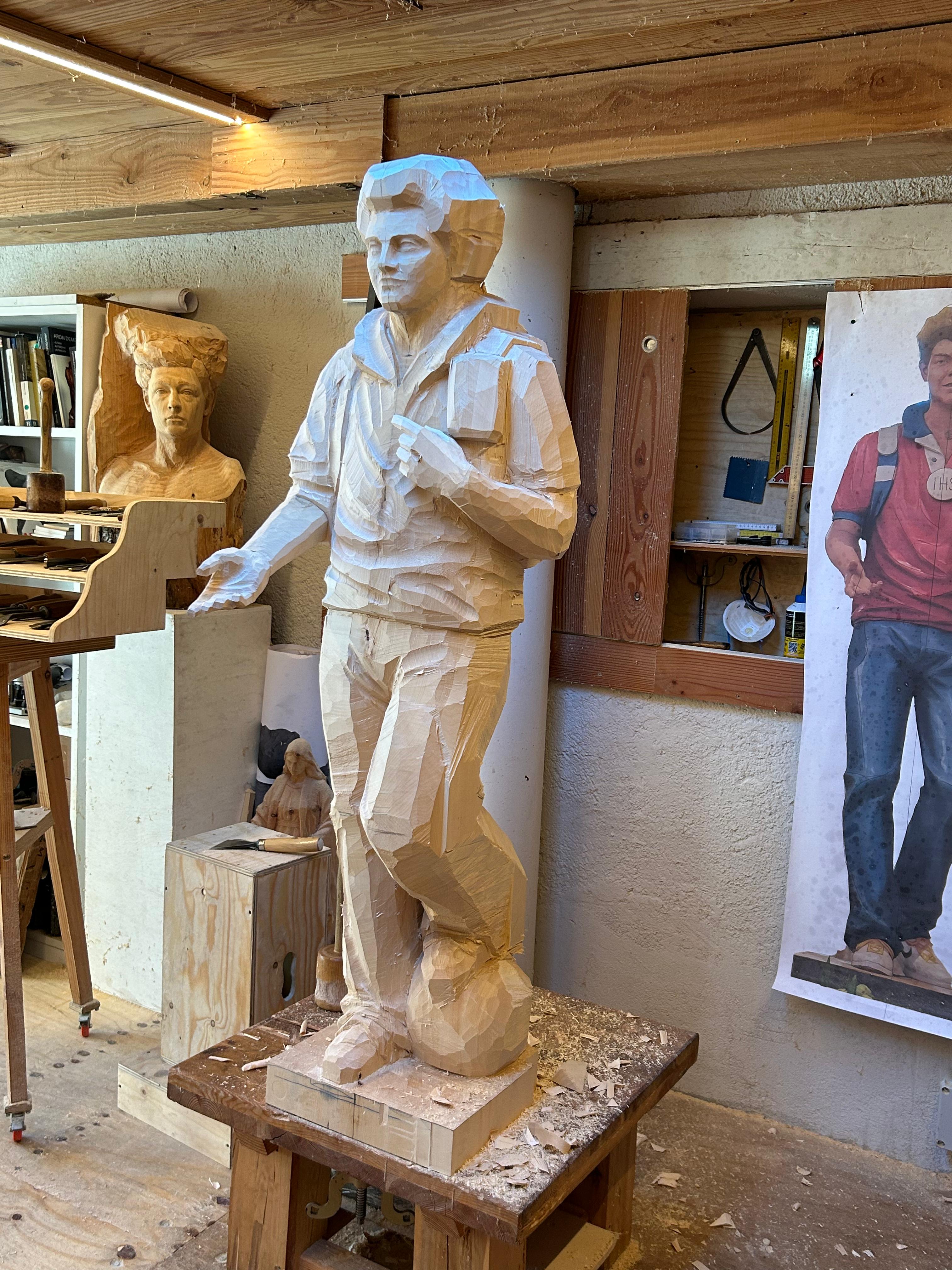 Blick auf die Carlo Acutis Statue aus einer anderen Perspektive während der Herstellung. Die Statue zeigt ihn mit einem Rucksack auf dem Rücken und einem Fuß auf einem Fußball. In einer Hand hält er ein Buch, in der anderen eine Kamera. Diese Statue wurde von der Schreinerei Ferdinand Stuflesser unter der Leitung von Robert Stuflesser gefertigt.