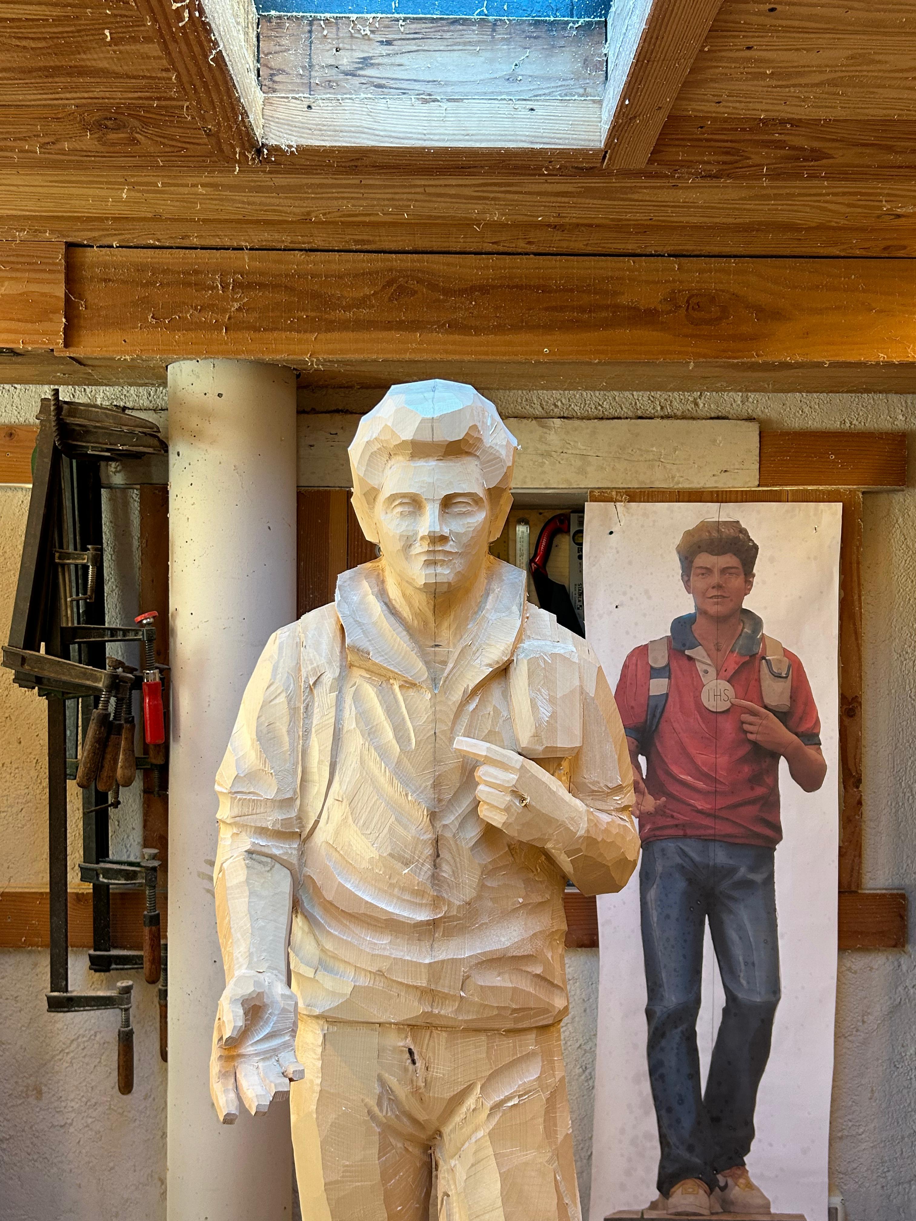 Detailaufnahme der Carlo Acutis Statue in der Herstellung, die ihn mit einem Rucksack auf dem Rücken zeigt. Eine Hand ist ausgestreckt, als würde er sie jemandem reichen. Diese Statue wurde von der Schreinerei Ferdinand Stuflesser unter der Leitung von Robert Stuflesser erschaffen.